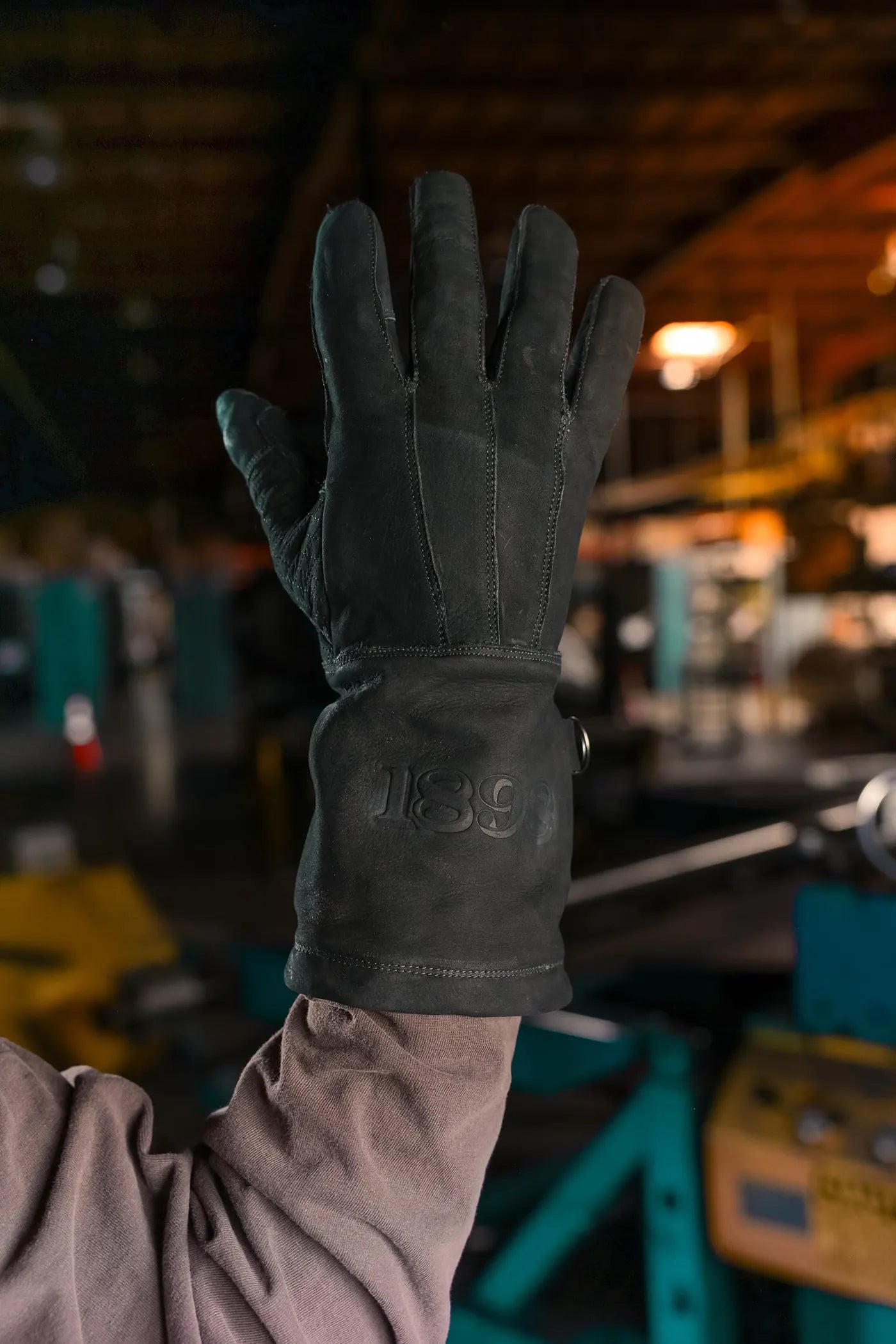 Black thread leather work glove held up in an industrial setting, featuring embossed 1898 brand logo on the wrist and textured leather surface Rated 4.70 – Black