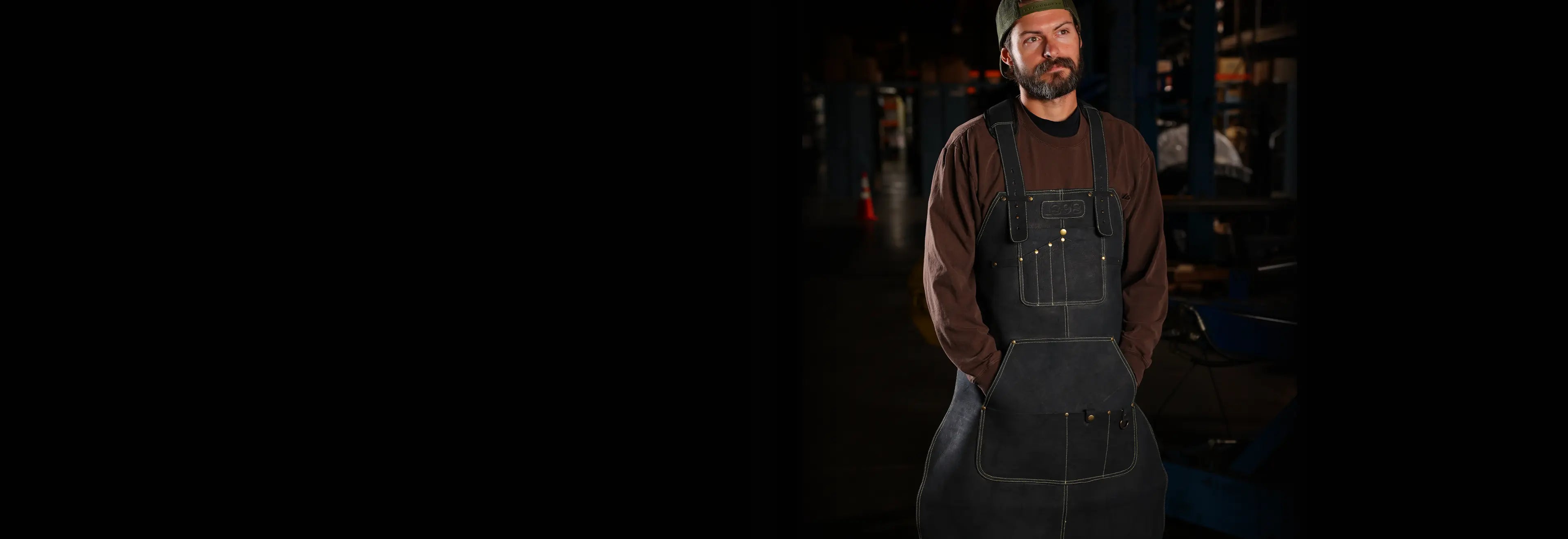 Craftsman wearing a black leather work apron with multiple pockets and reinforced stitching, standing in an industrial workshop with visible tools and machinery — designed for welders, metalworkers, and tradespeople who demand durability and function