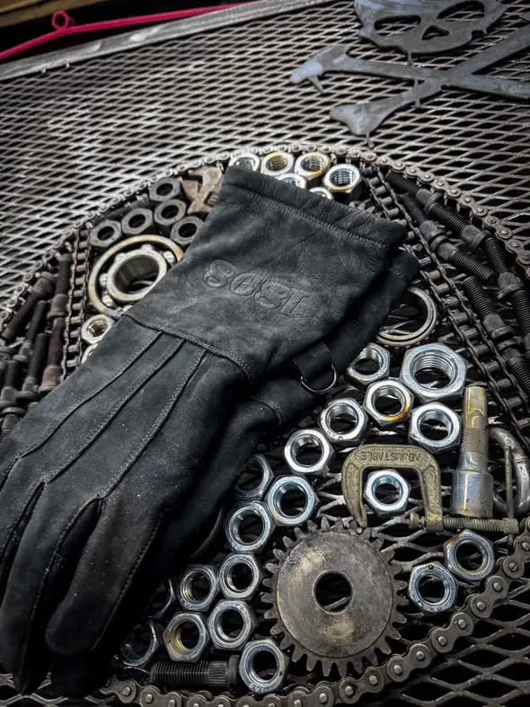 Black leather work glove laid on a circular arrangement of metal nuts, bolts, washers, and gears over a mesh surface, with a skull and crossbones design in the background — highlighting rugged design, mechanical utility, and craftsmanship – Rated 4.70 – Black