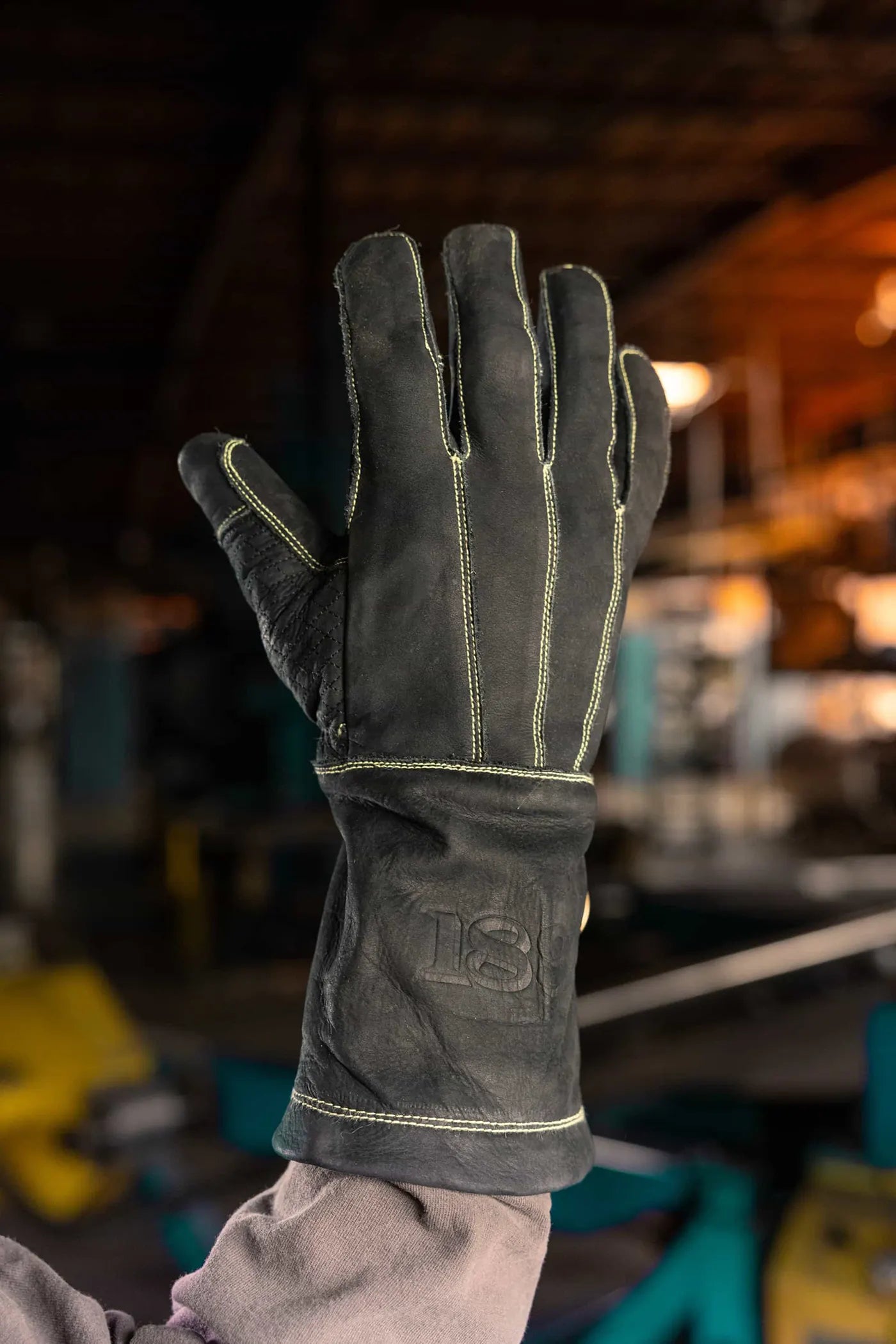 Leather work glove with gold stitching held up in an industrial setting, showing reinforced seams and “1898” embossed on the wrist Rated 4.70 – Gold