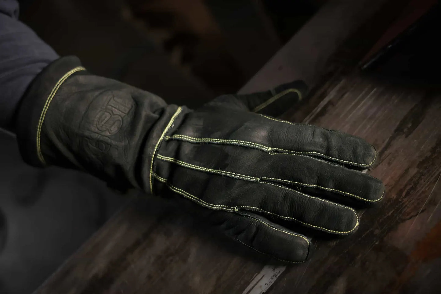 Close up of leather work glove with gold stitching resting on a wooden surface, showing detailed seams, craftsmanship, and faint 1898 logo on the wrist Rated 4.70 – Gold