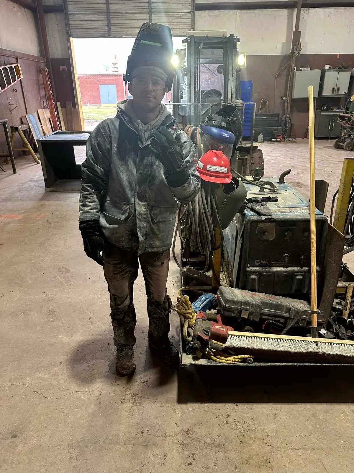 Person wearing gold-thread leather work gloves and welding gear in an industrial workshop, holding up three fingers with tools, ropes, and a forklift in the background — showcasing durability, fit, and real-world performance – Rated 4.60 – Gold