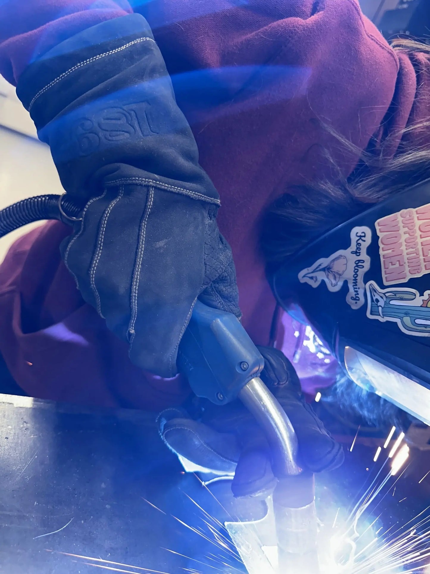 Welder wearing leather welding gloves with golden stitching and a helmet covered in stickers, actively welding with bright sparks and blue arc light — showcasing heat resistance, protection, and real-world performance – Rated 4.60 – Gold