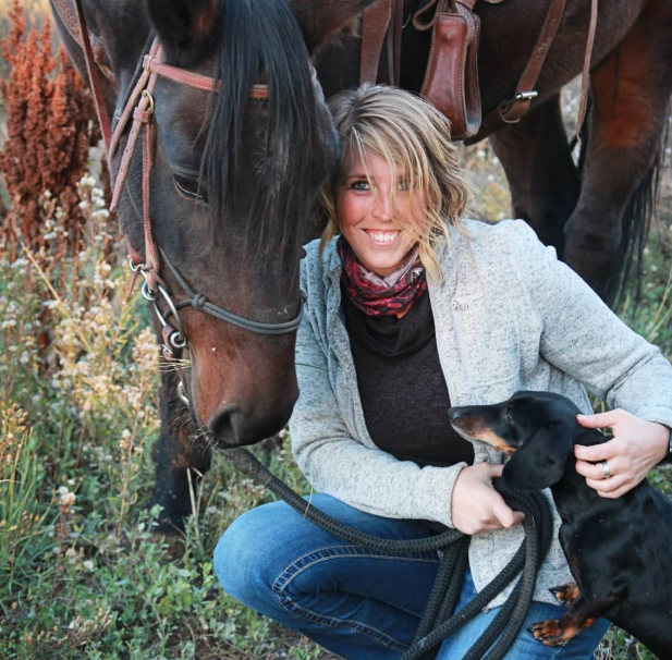 Metal artist Sarah of Ironmaden kneeling beside her horse with a dachshund on her lap