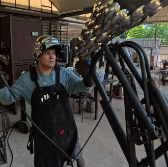 Sculptor Joaquin Soto in welding helmet and apron steadying a large steel sculpture in his outdoor shop