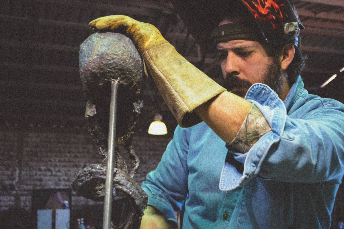 Sculptor David Madero in a flame-painted welding helmet steadying a skull sculpture with a leather gauntlet glove
