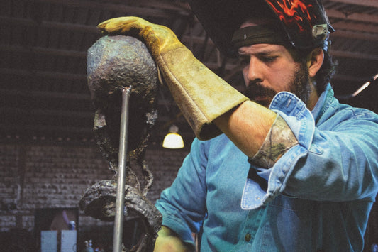 Sculptor David Madero in a flame-painted welding helmet steadying a skull sculpture with a leather gauntlet glove