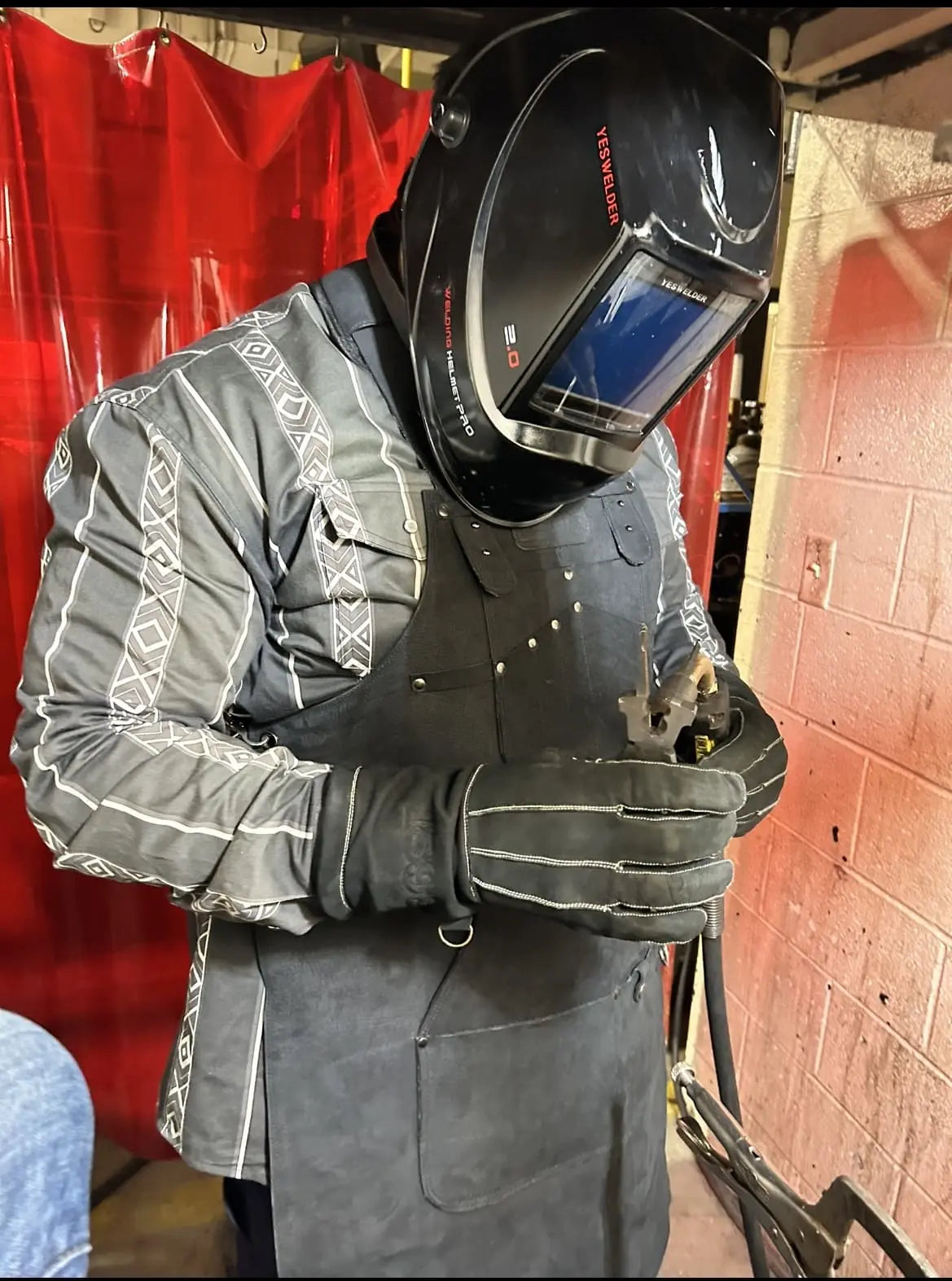 Person wearing a black leather work apron with white-thread detailing, full welding gear, and gloves while holding metal in an industrial workshop — showcasing protection, heat resistance, and durability in real-world metalworking – Rated 4.85 – Black