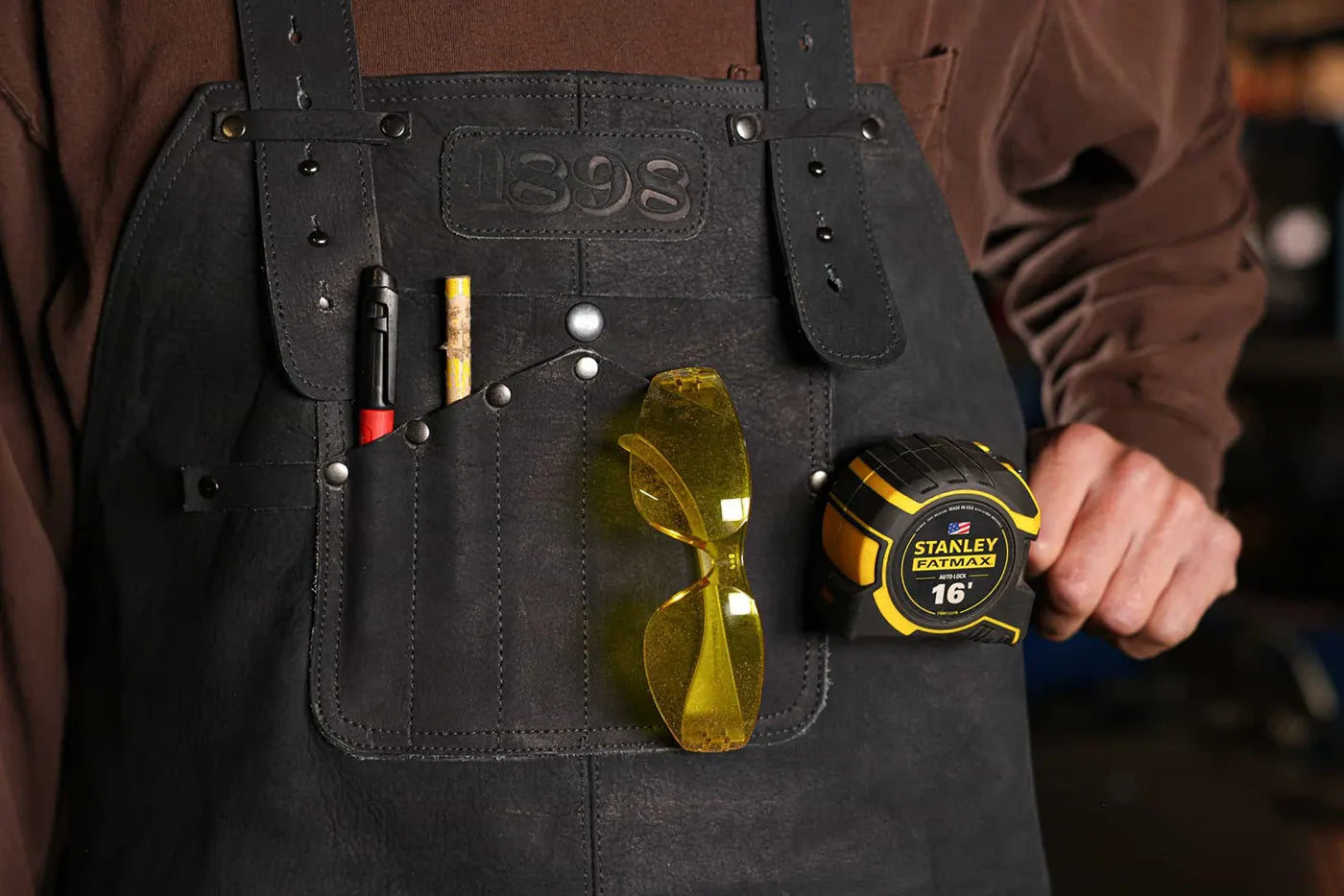 Front view of 1898 black leather welding apron with utility pockets holding pencil, pen, safety glasses, and Stanley FatMax tape measure – Black