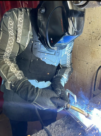 Person wearing a black leather work apron with black stitching, welding helmet, and gloves while actively welding in an industrial workshop — sparks and blue arc light highlight protection, heat resistance, and premium craftsmanship – Rated 4.85 – Black
