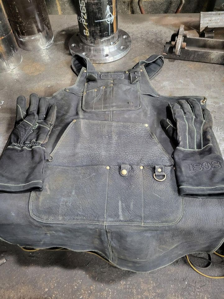 Leather work apron with gold stitching, brass hardware, and multiple pockets laid on a metal surface with a pair of gloves embroidered with “1898” — highlighting premium craftsmanship, reinforced build, and trade-ready design – Rated 4.85 – Gold