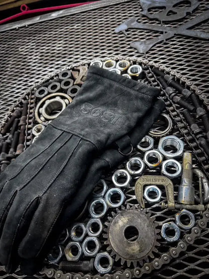 Black leather work glove laid on a circular arrangement of metal nuts, bolts, washers, and gears over a mesh surface, with a skull and crossbones design in the background — highlighting rugged design, mechanical utility, and craftsmanship – Rated 4.70 – Black