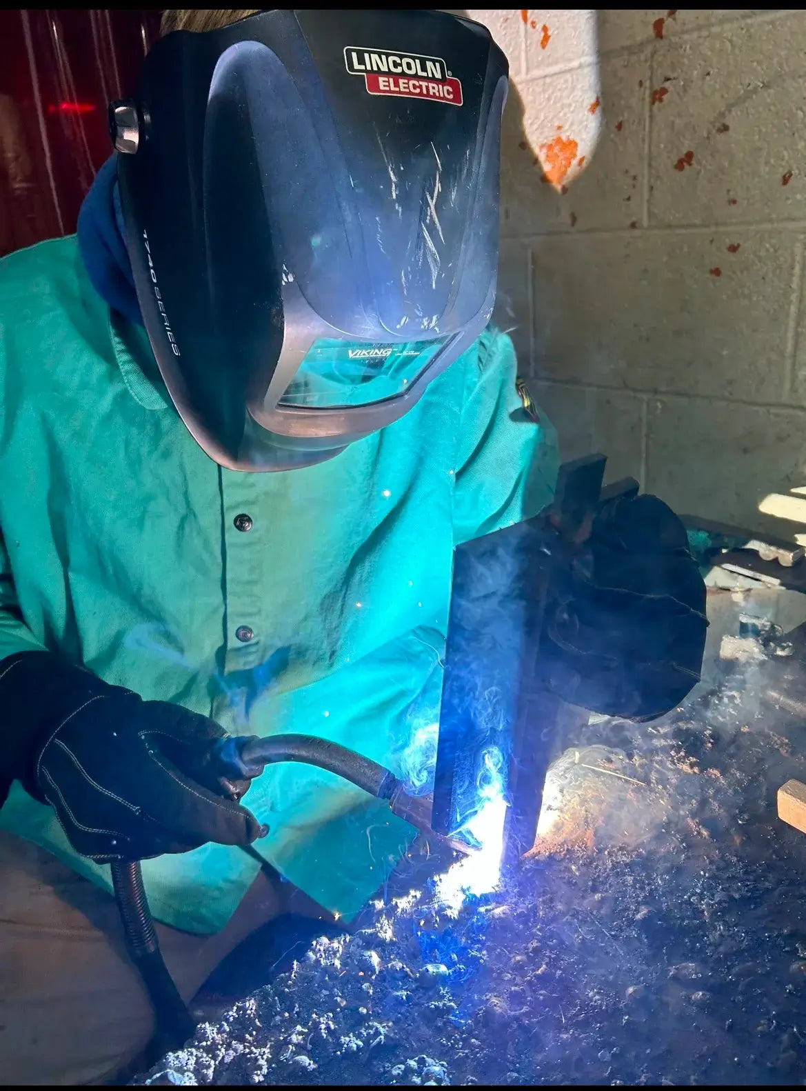 A student welder wearing a Lincoln Electric helmet and teal safety jacket focuses on a live weld during the 1898 collaboration with Warren County Career Center.

